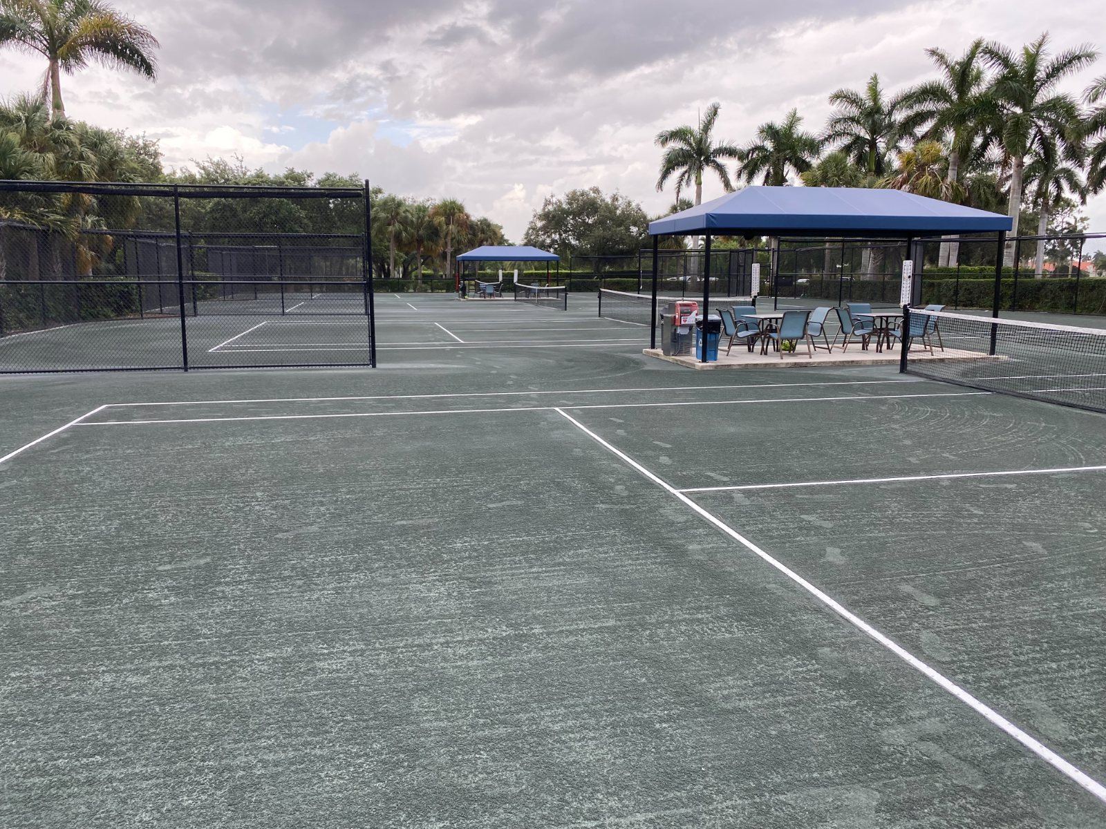 Multiple tennis courts with blue shade structures, seating areas, and palm trees under cloudy sky