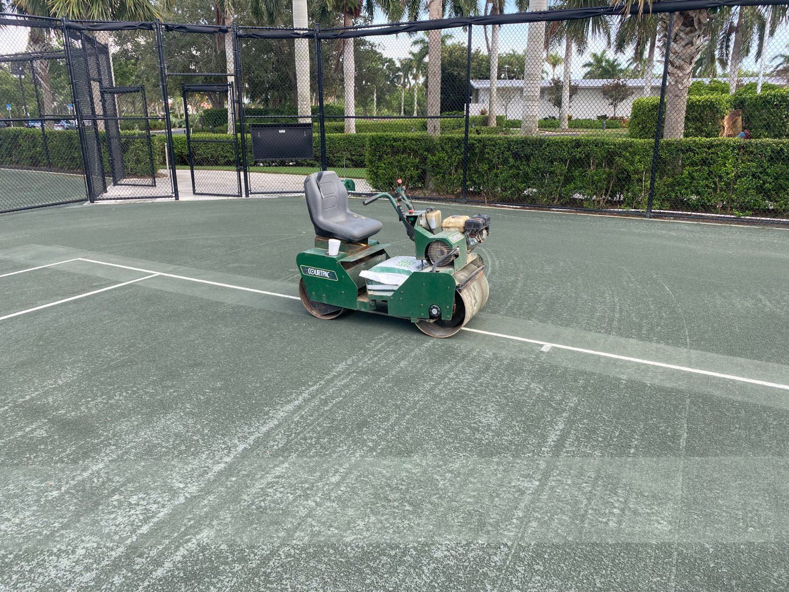 Green riding power trowel machine on tennis court with fencing and palms in background