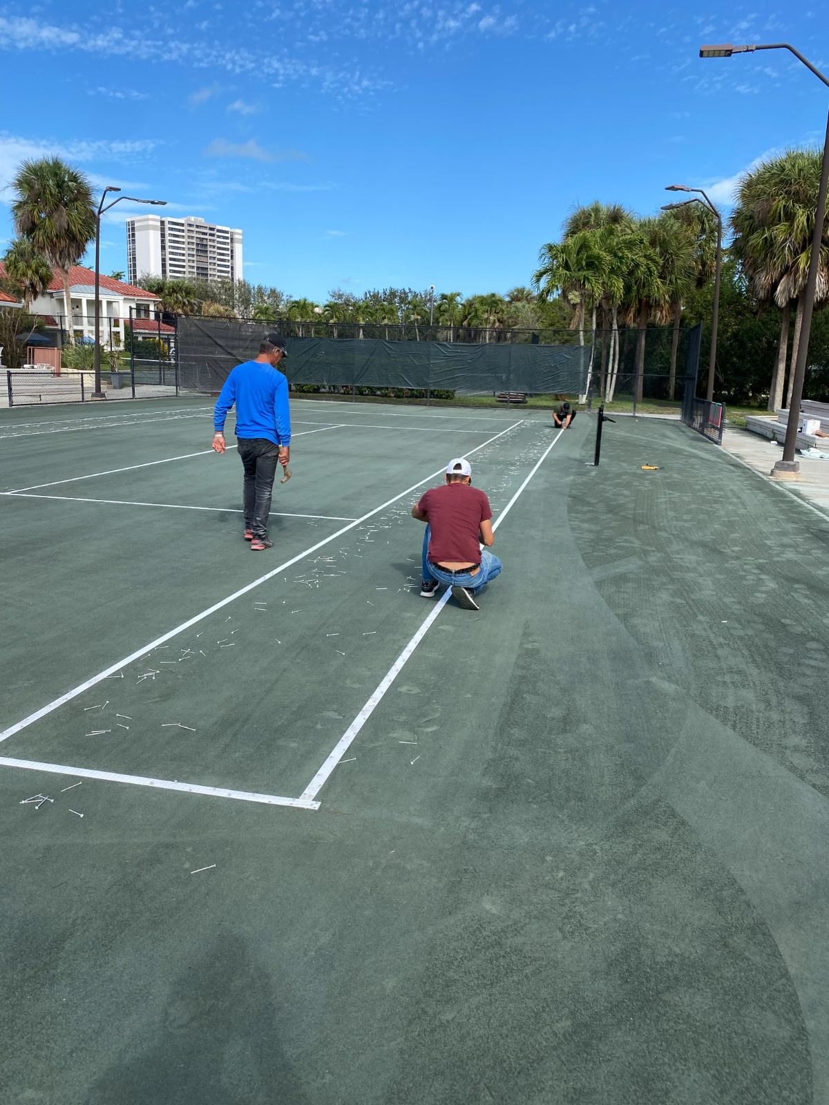 Three people playing tennis on outdoor courts with palm trees and tall buildings in background