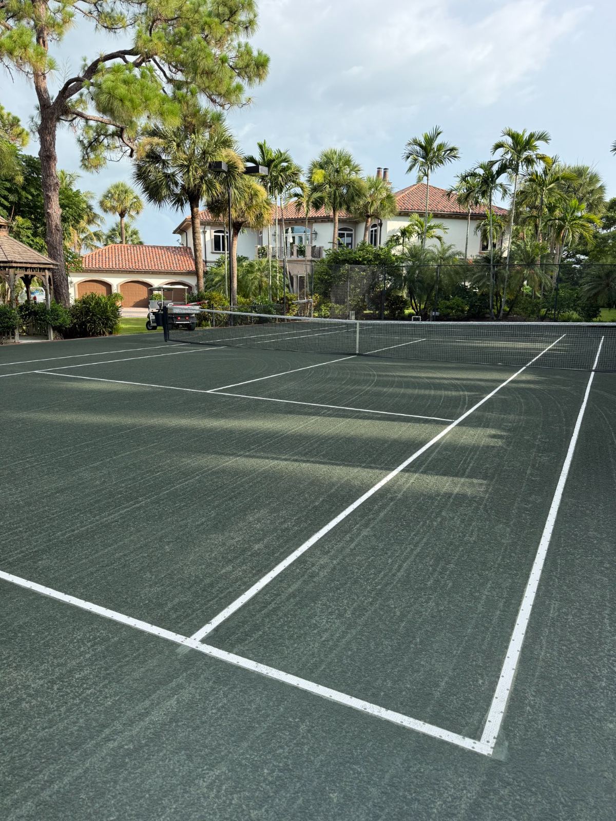Two professional tennis courts with white lines and net, luxury homes with palm trees in background