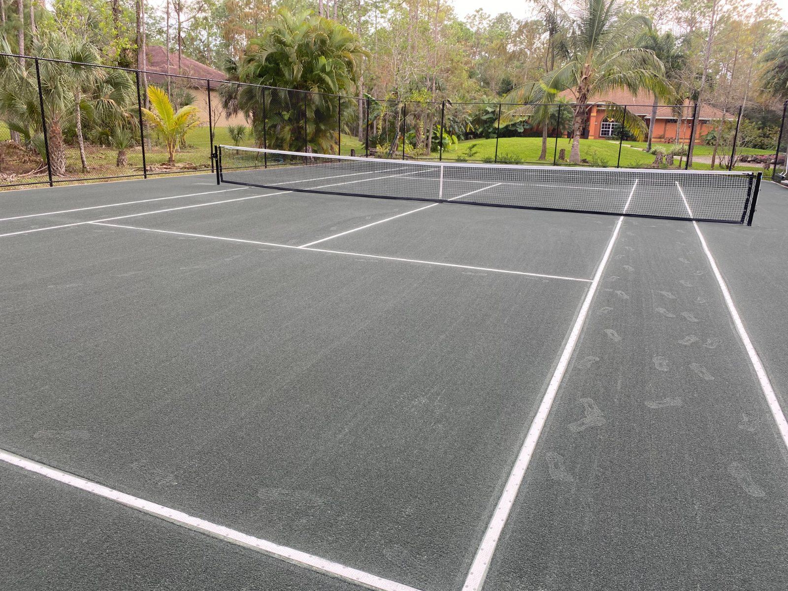 Two tennis courts with white line markings and black nets in a gated facility surrounded by palm trees and green landscape